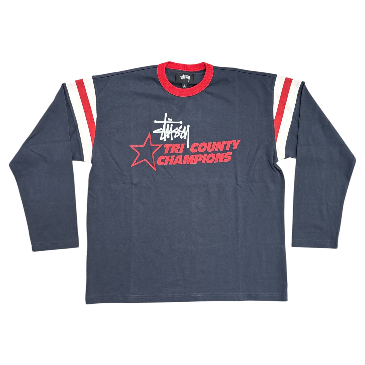 A Stussy long-sleeve ringer t-shirt in dark navy blue, featuring a red crew neck collar, red and white shoulder stripes, and a red and white Tri-County Champions graphic on the chest.
